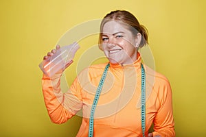 Happy smiley young sporty woman drinking water with measuring green tape on her neck over yellow background