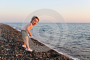 Happy smiling 2 years boy standing on the pebbles beach at sunset