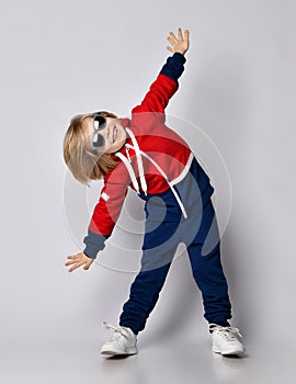 Happy smiling active blond kid boy in blue and red hoodie, pants and sunglasses does gym exercises