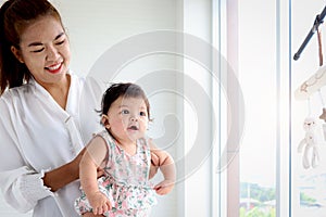 Happy smiling adorable six months baby girl standing next to the window in embrace of mother arms, mom holding her sweet little