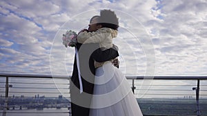 Happy smiling African American bride hugging groom standing on bridge outdoors. Camera moves around loving affectionate