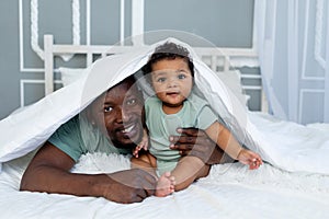 Happy smiling African american dad with baby son on the bed at home cuddling under the blanket, happy family, father`s day photo