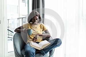 Happy smiling african american woman drink coffee sitting on sofa and reading book at home