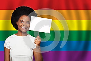 Happy smiling african american woman holding blank white sign on LGBT flag background