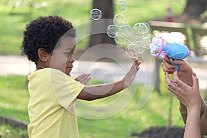 Happy smiling African boy with black curly hair playing with soap bubbles making from blowing bubble gun toy at green garden.