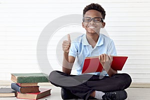 Happy smiling African boy with glasses reading book and giving thumb up while sitting on floor in white wall room. Portrait of