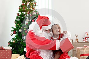Happy smiling African child girl baby sitting on Santa Claus lap with decorative Christmas tree as background, cute kid open