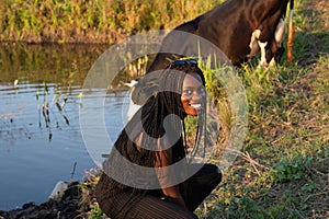 Happy smiling african girl in black clothes crouched and looking at the camera, cow drinks water from the lake on the