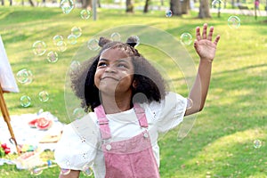 Happy smiling African girl with black curly hair blowing and playing with soap bubbles at green garden. Cute child has fun with