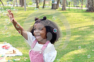 Happy smiling African girl with black curly hair blowing and playing with soap bubbles at green garden. Cute child has fun with