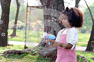 Happy smiling African girl with black curly hair making soap bubbles with blowing bubble gun toy at green garden. Kid spending
