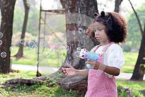 Happy smiling African girl with black curly hair making soap bubbles with blowing bubble gun toy at green garden. Kid spending