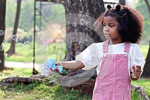 Happy smiling African girl with black curly hair making soap bubbles with blowing bubble gun toy at green garden. Kid spending