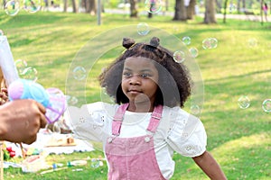 Happy smiling African girl with black curly hair playing with soap bubbles making from blowing bubble gun toy at green garden.