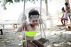 Happy smiling African girl with black curly hair wearing brightly colored swimsuit, sitting on swing at beach outdoor, beautiful