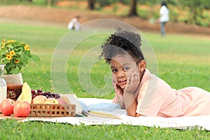 Happy smiling African little girl with black curly hair lying down on mat on green grass. Cute kid spending time outdoor in green