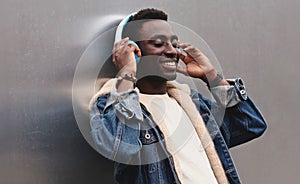 Happy smiling african man in wireless headphones listening to music on city street over gray metal wall