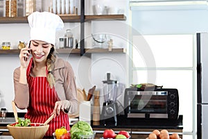 Happy smiling Asian beautiful woman wear apron and chef hat, talking on mobile phone during prepare fresh vegetables salad at