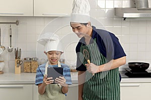 Happy smiling Asian boy kid wears apron and chef hat, holds digital tablet for searching recipes online Internet, son and father