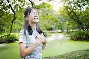 Happy smiling asian child girl standing in green nature,holding hands on heart,female teenage touching her chest,enjoy breathing