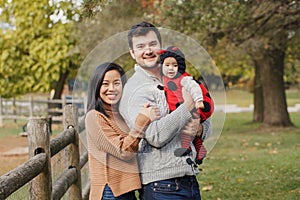 Happy smiling Asian Chinese mother and Caucasian father dad with baby girl in ladybug costume. Family in autumn fall park outdoors