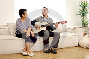 Happy smiling Asian elderly couple relaxing and playing guitar while sitting on sofa in the living room, romantic senior mature