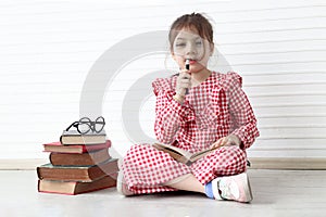 Happy smiling Asian girl holding magnifying glass while sitting on floor with book stack in white wall room. Cute child with pile