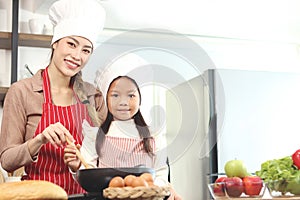 Happy smiling Asian mother and daughter wearing apron and chef hat cooking tasty fried food meal with pan at kitchen with fresh