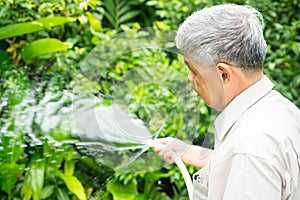 A happy and smiling Asian old elderly man is watering plants and flowers for a hobby after retirement in a home. Concept of a