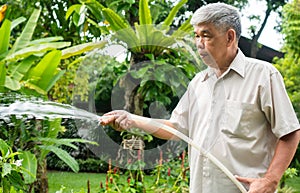 A happy and smiling Asian old elderly man is watering plants and flowers for a hobby after retirement in a home. Concept of a