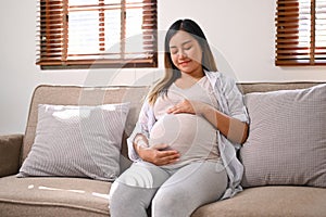 Happy Asian pregnant woman relaxes on the sofa in her living room, touching belly with love