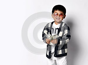 Happy smiling asian rich kid boy millionaire in modern sunglasses stands holding fan of dollars cash money. Lucky winner