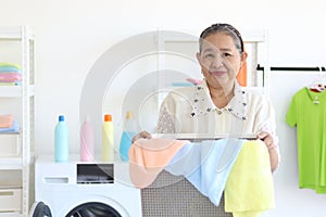 Happy smiling Asian senior elderly woman housewife carrying clothes basket for doing laundry at laundry room with washing machine