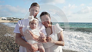 Happy smiling Asian woman and Caucasian man posing with cute baby girl on turquoise Mediterranean sea beach. Positive