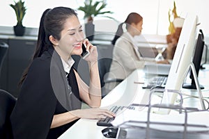 Happy smiling Asian woman officer sitting, talking on mobile phone at office desk with blurred busy colleague background, modern