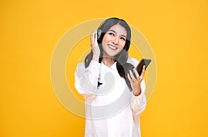 Happy smiling Asian woman wearing wireless headphones listening to music with smartphone on bright yellow background