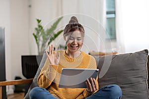 Happy smiling asian young woman in yellow sweater with tablet pc computer having video call and waving hand at home