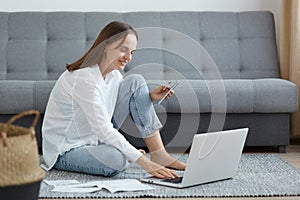 Happy smiling attractive woman wearing white shirt and jeans sitting on floor near sofa, using laptop, holding credit card and