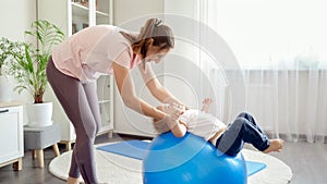 Happy smiling baby boy rolling on blue fitness ball with smiling mother doing exercises at home