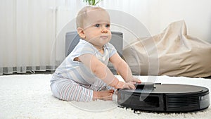 Happy smiling baby boy sitting on carpet and playing with robot vacuum cleaner. Concept of hygiene, household gadgets