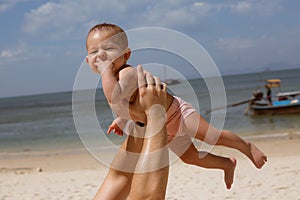 Happy smiling baby in dad arms. On a tropical beach. Sunny day, father throws up infant toddler, hold on hands. Child enjoyed