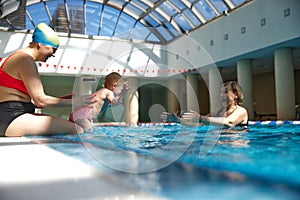 Happy, smiling baby girl, child having fun in indoor swimming pool with her mother and grandmother. Jumping into water