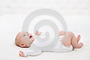 Happy Smiling Baby in White Cotton Onesies in Bed lying on Back Side View. Laughing Infant Child in babies Bodysuit play