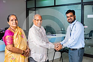 Happy smiling banker greeting by shaking hand to senior couple at bank - concept of financial support, banking adviser