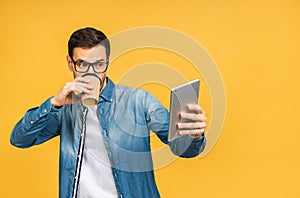 Happy smiling bearded man using digital tablet, reading media news, isolated over yellow background. Drinking cop of coffee