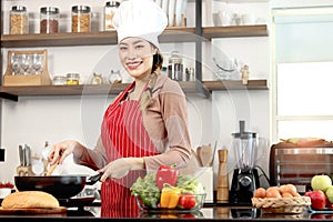 Happy smiling beautiful Asian woman wearing apron and chef hat, holding turner for cooking tasty fried food meal with pan at