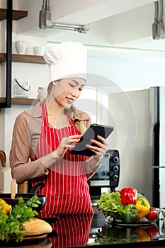 Happy smiling beautiful Asian woman wears apron and chef hat, holding and looking at digital tablet for searching tasty food meal