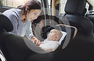 Happy Smiling Beautiful Asian young mother putting her baby son into car seat and fasten seat belts in the car