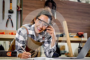 Happy smiling beautiful carpenter woman wearing safety glasses goggles and apron taking to customer by using mobile phone, female