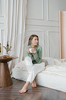 Happy smiling beautiful European woman enjoys the morning and inhales the aroma of a cup of coffee in her bedroom. Enjoying free a